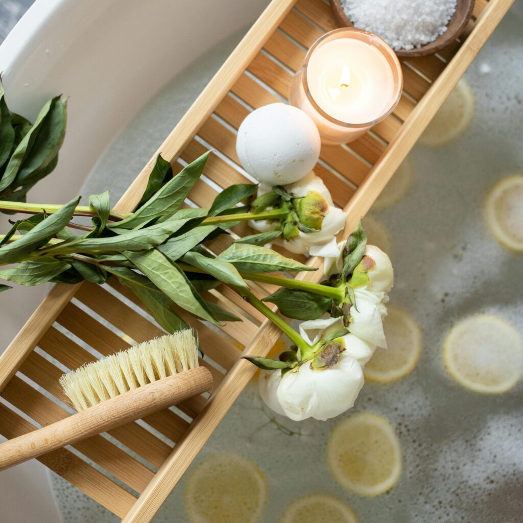 Top view of wooden tray with bath bomb and burning candle placed near salt and flowers on tub with lemon slices on water