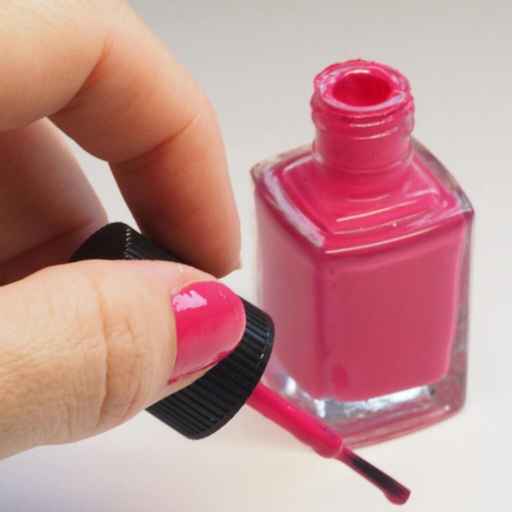 Female hand applying bright pink nail polish from open bottle, minimalistic style.